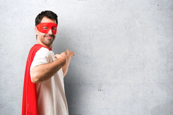 Superhero man with mask and red cape pointing back with the index finger presenting a product from behind on textured grey background