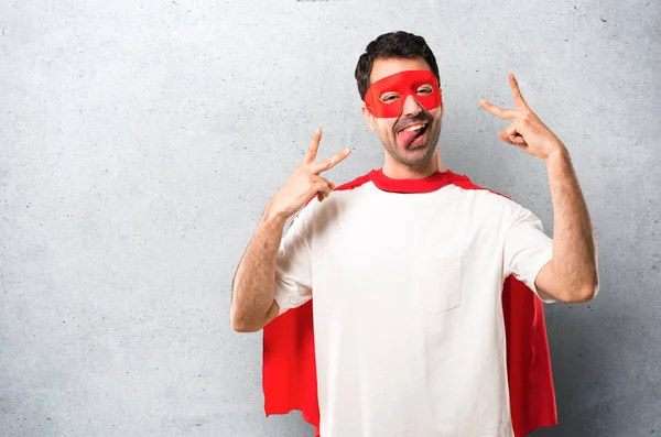質感の灰色の背景に面白い顔を持つカメラで舌を示すマスクと赤のケープとスーパー ヒーローの男 — ストック写真