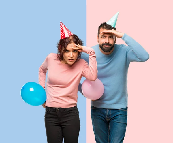 couple with balloons and birthday hats looking far away with hand to look something on pink and blue background