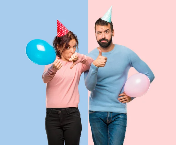 couple with balloons and birthday hats making good-bad sign. Undecided between yes or not on pink and blue background