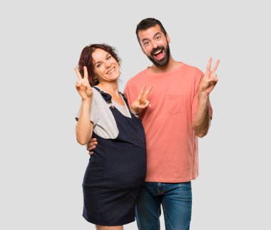Çift gülümsüyor ve her ikisi de zafer işaretiyle gösterilen hamile kadın ile izole gri arka plan üzerinde eller