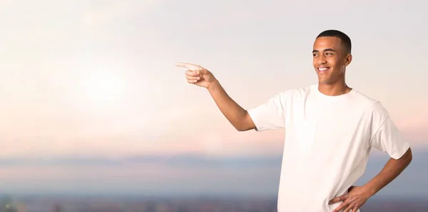 Young african american man pointing finger to the side and presenting a product while smiling in a confident pose on sunset background