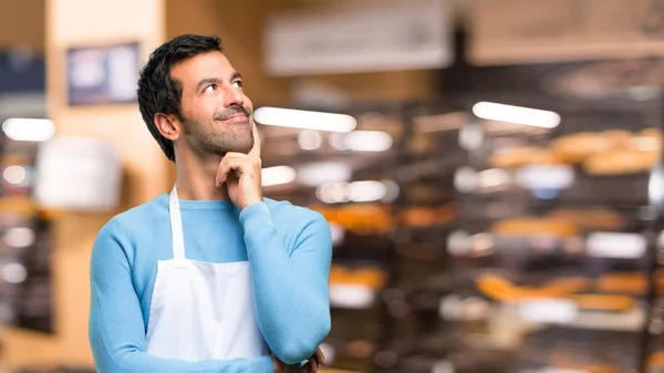 Man wearing an apron standing and thinking an idea while looking up in a bakery