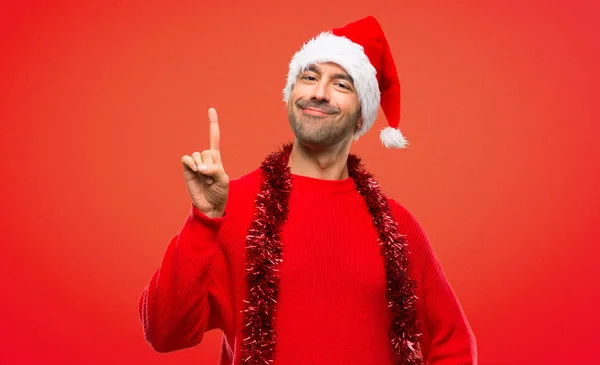 Man with red clothes celebrating the Christmas holidays showing and lifting a finger in sign of the best on red background