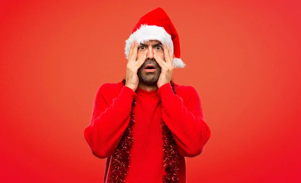 穿着红色衣服庆祝圣诞节假期的男子惊讶地用双手捂住脸 同时用手指在红色背景上看 — 图库照片