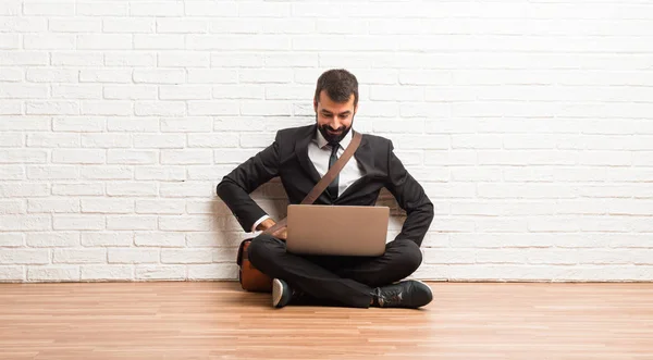Businessman with his laptop sitting on the floor posing with arms at hip and laughing looking to the front