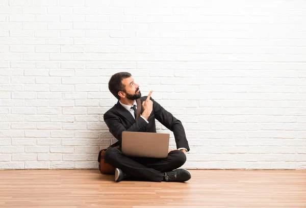 Businessman with his laptop sitting on the floor pointing back with the index finger