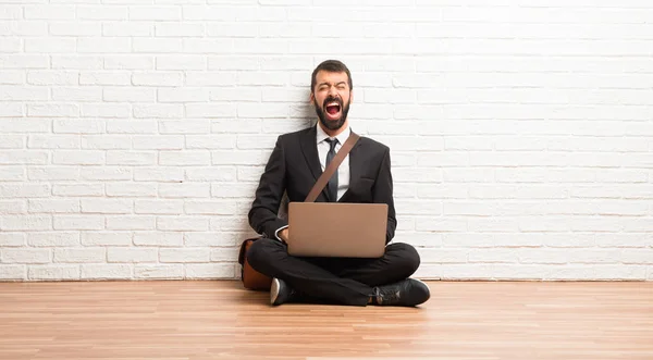 Businessman with his laptop sitting on the floor shouting to the front with mouth wide open
