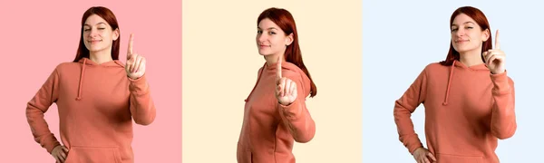 一套年轻的红头发的女孩与粉红色的运动衫显示和举起手指在五颜六色的背景上的最佳标志 — 图库照片