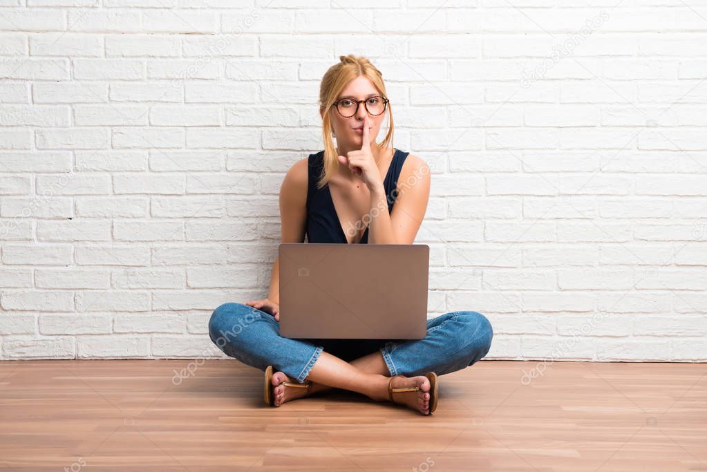 Blonde girl sitting on the floor with her laptop showing a sign of closing mouth and silence gesture on white brick wall background