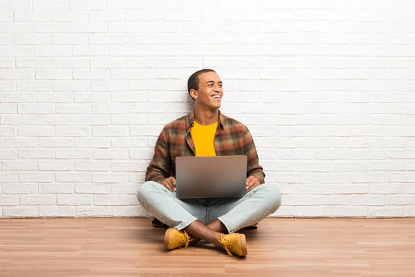 非洲裔美国人坐在地板上 他的笔记本电脑摆姿势与手臂在臀部和笑 — 图库照片