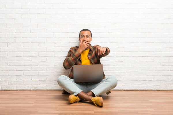 African american man sitting on the floor with his laptop pointing with finger at someone and laughing