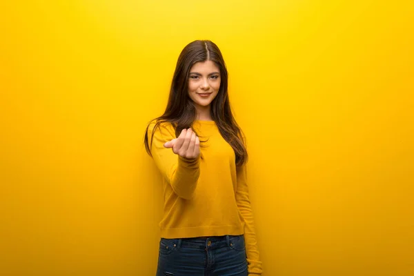Teenager girl on vibrant yellow background inviting to come with hand. Happy that you came