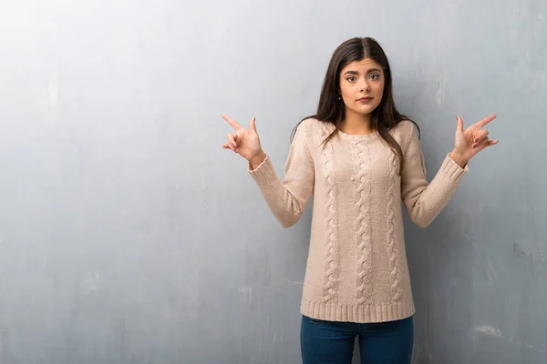 Teenager girl with sweater on a vintage wall pointing to the laterals having doubts