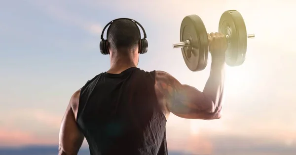 Strong handsome sport man making weightlifting, listening music and  exercising the shoulders on sunset background