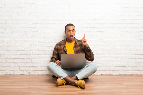 African american man sitting on the floor with his laptop thinking an idea pointing the finger up