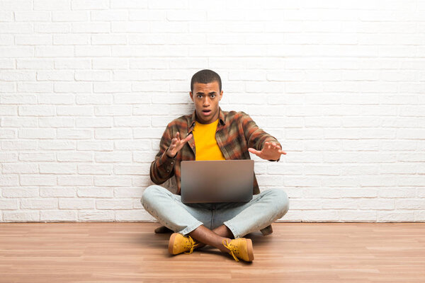 African american man sitting on the floor with his laptop is a little bit nervous and scared stretching hands to the front