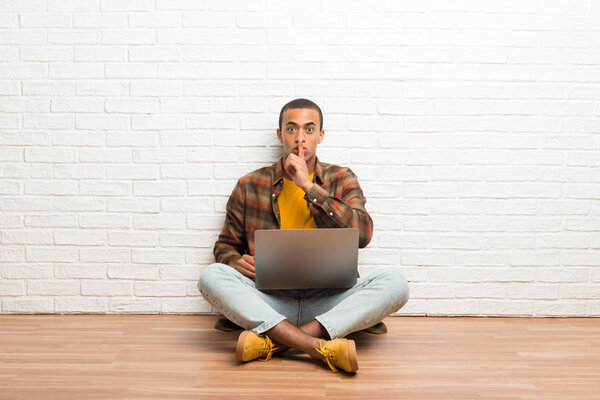 African american man sitting on the floor with his laptop showing a sign of silence gesture putting finger in mouth