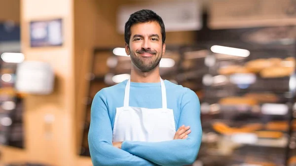 Man wearing an apron keeping the arms crossed in frontal position. Confident expression in a bakery