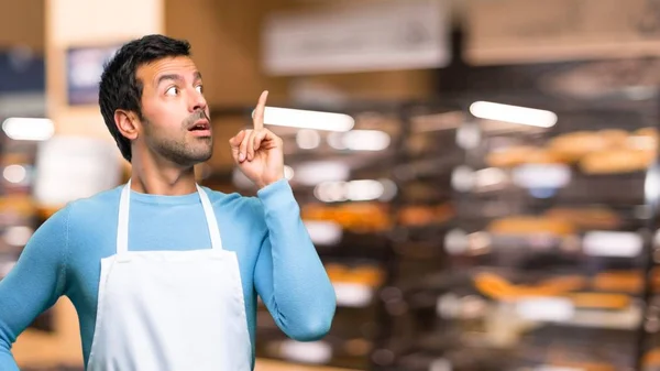 Man wearing an apron standing and thinking an idea pointing the finger up in a bakery