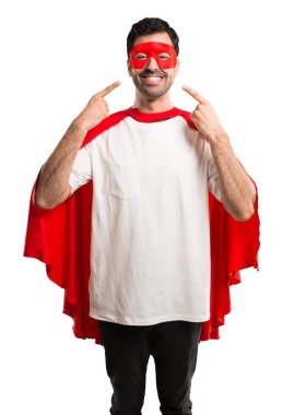 Süper kahraman maske ve kırmızı pelerin ağız ve yüz parmak ile izole beyaz zemin üzerine işaret ederken mutlu ve keyifli bir ifade ile gülümseyen adam