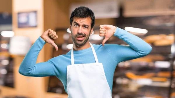 Man wearing an apron showing thumb down with both hands. Negative expression in a bakery