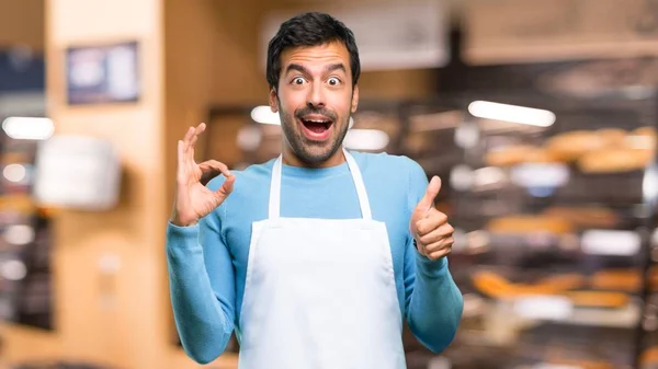 Man wearing an apron showing an ok sign with fingers and giving a thumb up gesture with the other hand in a bakery