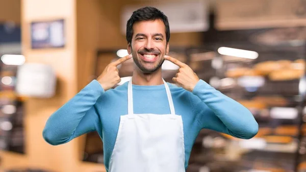 Man wearing an apron smiling with a happy and pleasant expression while pointing mouth and face with fingers in a bakery