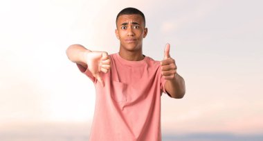 İyi-kötü işaret yapma adam koyu tenli. Kararsız kişi Evet arasında veya değil günbatımı arka