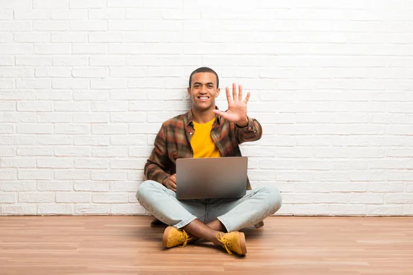 African american man sitting on the floor with his laptop counting five with fingers