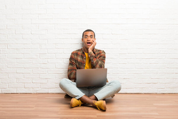 African american man sitting on the floor with his laptop surprised and shocked while looking right