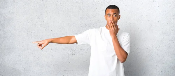 Young african american man pointing finger to the side and presenting a product with a surprised face while covering mouth