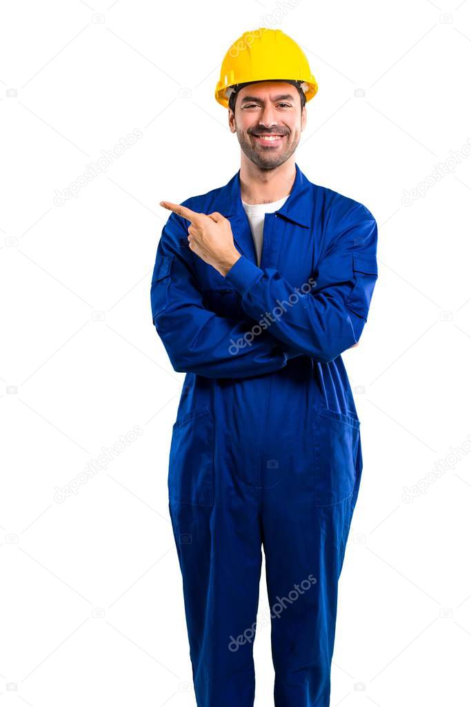 Young workman with helmet pointing to the side with a finger to present a product or an idea while looking forward smiling on isolated white background