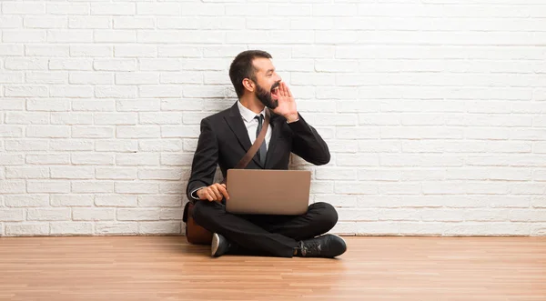 Businessman with his laptop sitting on the floor shouting with mouth wide open to the lateral