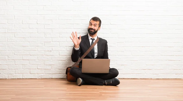Businessman with his laptop sitting on the floor saluting with hand with happy expression
