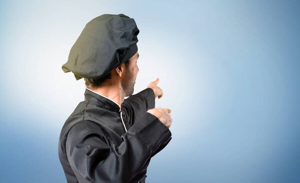 Chef man In black uniform pointing back with the index finger presenting a product from behind on blue background