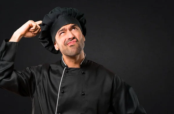 Chef man In black uniform having doubts and with confuse face expression while scratching head on black background