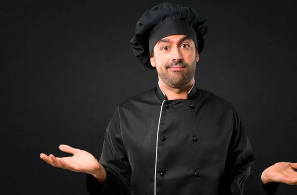 Chef man In black uniform having doubts and with confuse face expression while raising hands and shoulders Uncertain concept on black background