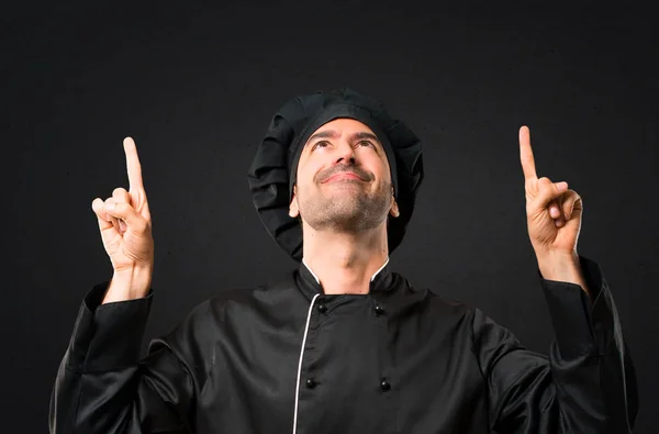Chef man In black uniform pointing with the index finger a great idea and looking up on black background