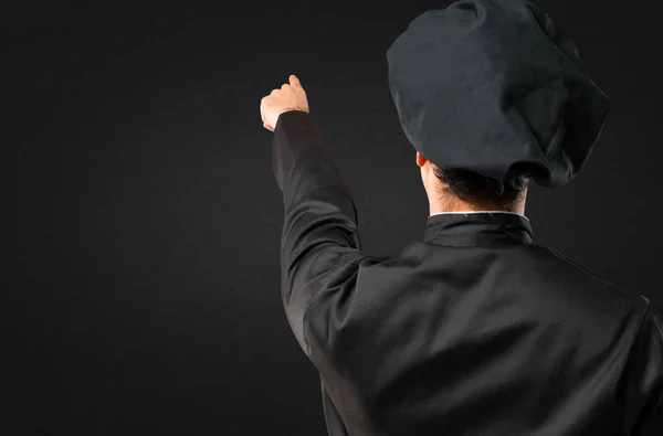 Chef man In black uniform pointing back with the index finger on black background