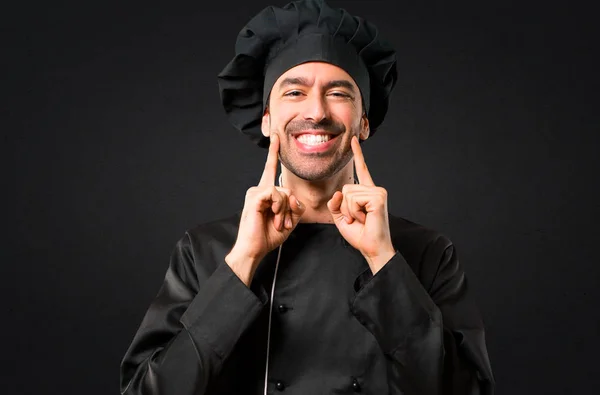 Chef man In black uniform smiling with a happy and pleasant expression while pointing mouth and face with fingers on black background