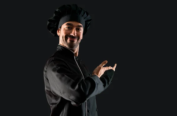 Chef man In black uniform presenting and inviting to come with hand on black background
