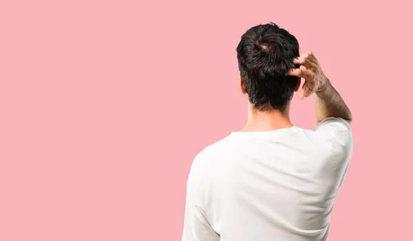 Young man with white shirt on back position looking back while scratching head on isolated pink background