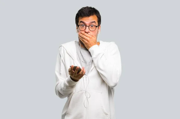 Man with glasses and listening music covering mouth with both hands for saying something inappropriate. Can not speak on grey background