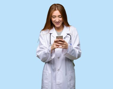 Kızıl saçlı doktor kadın izole mavi arka plan üzerinde mobil ileti gönderme