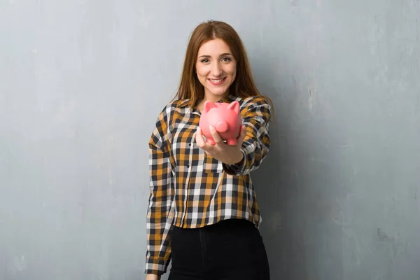 Joven Pelirroja Sobre Grunge Pared Sosteniendo Piggybank —  Fotos de Stock