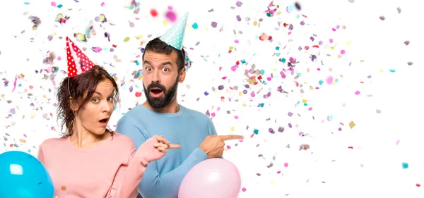 Pareja Con Globos Sombreros Cumpleaños Señalando Dedo Lado Con Una —  Fotos de Stock