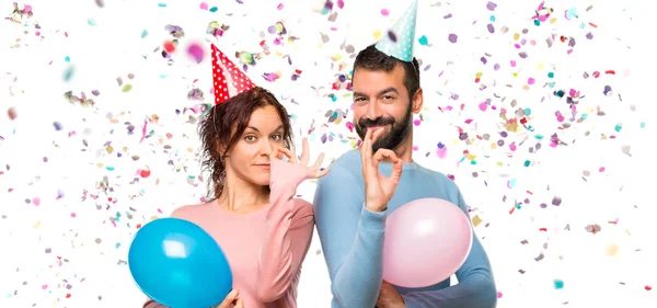 couple with balloons and birthday hats showing an ok sign with fingers with confetti in a party