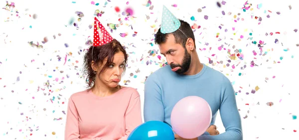 couple with balloons and birthday hats with sad and depressed expression. Serious gesture with confetti in a party