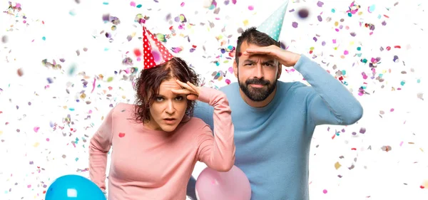 couple with balloons and birthday hats looking far away with hand to look something with confetti in a party